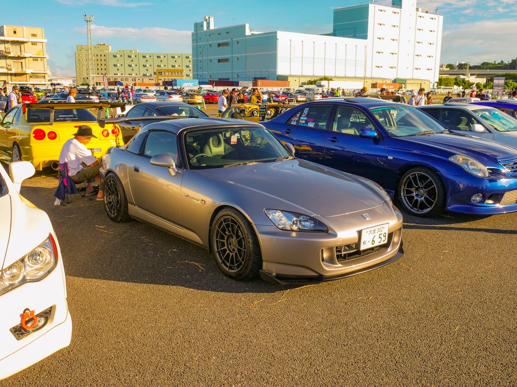 A lively car meet with JDM models including a silver Honda S2000 and other sports cars under the sunny sky.