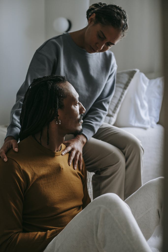 Attentive loving African American female supporting and touching shoulders of stressed frustrated husband while sitting in light bedroom