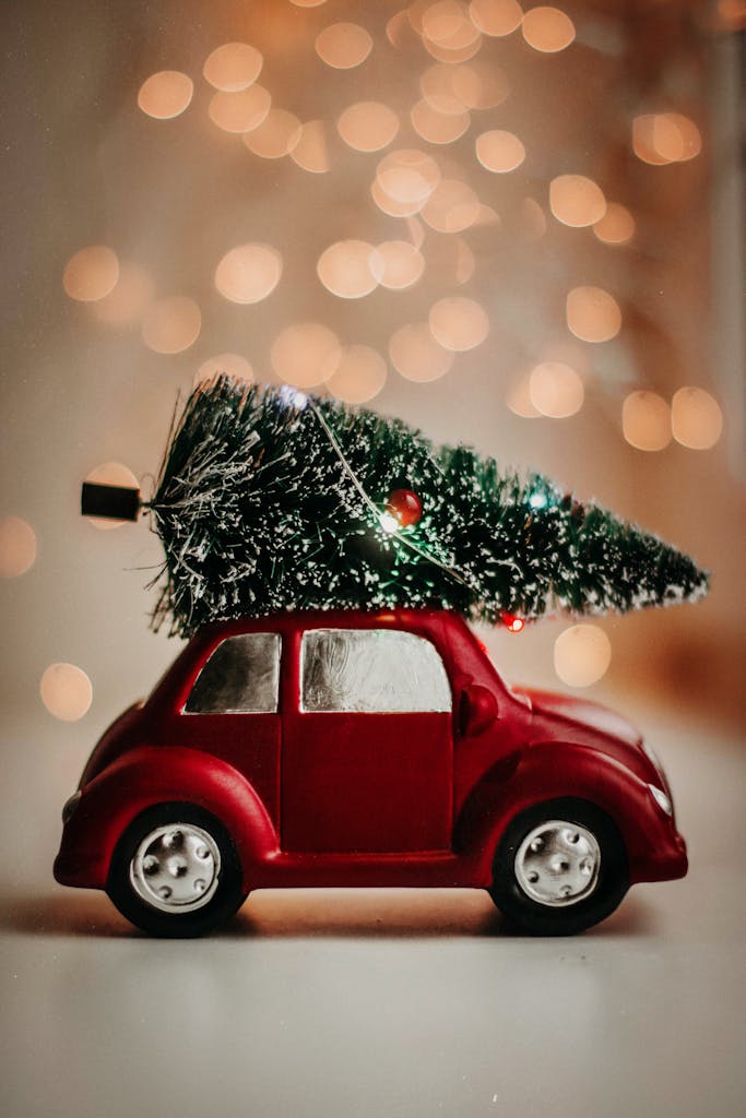 A red toy car carries a Christmas tree, set against a warm bokeh background, celebrating the festive season.