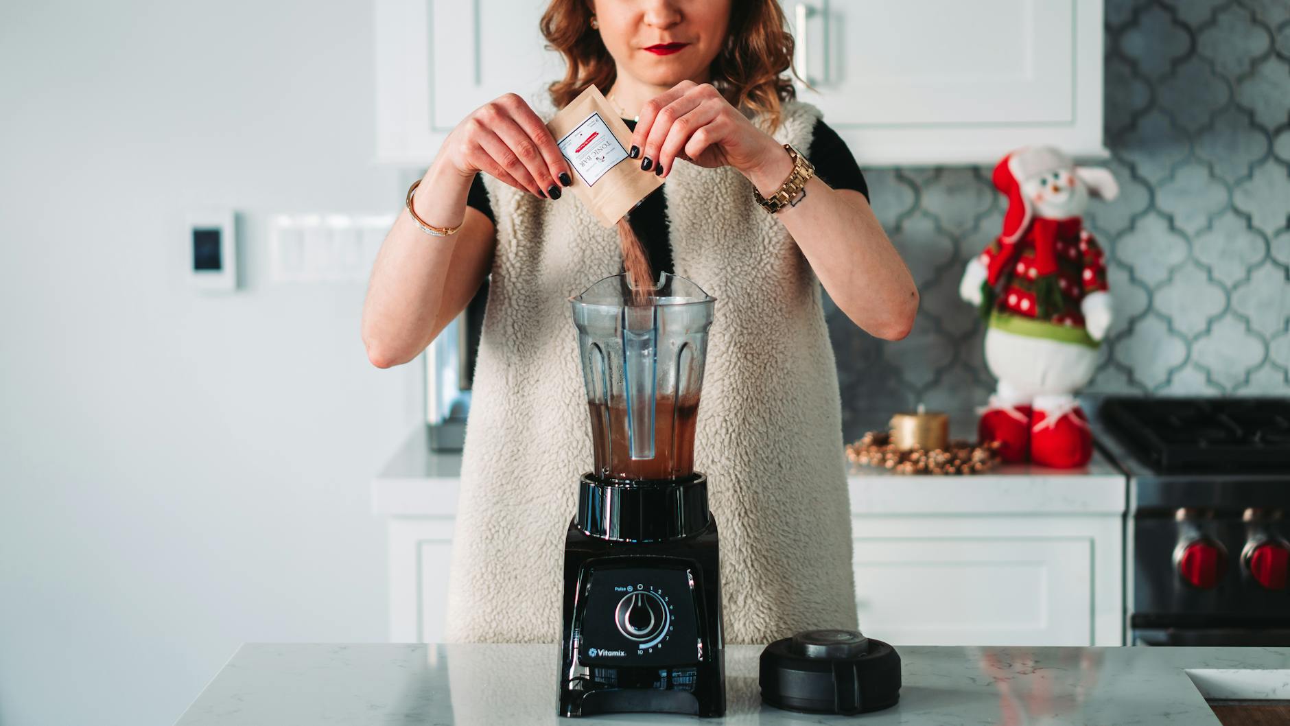 Vitamix Blender On Kitchen Countertop With Ingredients Like Kale, Frozen Fruit, And Nuts